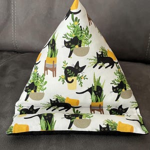 May include: A white triangular pillow with a black cat and plant pattern. The cats are black with yellow eyes, and the plants are green with yellow flowers. The pillow has a black border and is resting on a gray surface.