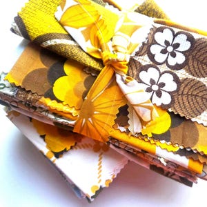 May include: A stack of vintage fabric swatches in shades of yellow, brown, and white, tied with a matching fabric ribbon. The fabrics feature floral and geometric patterns, with a retro aesthetic. The edges of the fabric are unfinished.