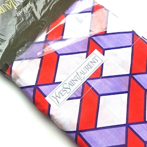 May include: A folded textile featuring a geometric pattern of white, red and purple cubes. The fabric is encased in clear plastic packaging with a white label displaying the text Yves Saint Laurent in black lettering and a black header.