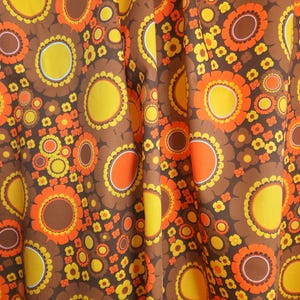 May include: A fabric featuring a dense, repeating pattern of large and small circular flower motifs in shades of brown, bright orange, and yellow. The design includes layered petals and solid centres, creating a continuous, vibrant floral print.