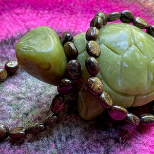 Könnte beinhalten: Ein braunes und goldenes Edelstein-Armband mit ovalen Perlen, das um eine grüne Jade-Schildkrötenfigur gewickelt ist. Das Armband liegt auf einer rosa-grünen, flauschigen Oberfläche.