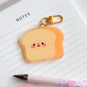 Könnte beinhalten: Ein klarer Acryl-Schlüsselanhänger in Form einer Brotscheibe mit einem lächelnden Gesicht und goldfarbener Hardware. Der Anhänger liegt auf einem linierten Notizblock mit dem Wort "NOTES" und wird von rosa Stiften begleitet.