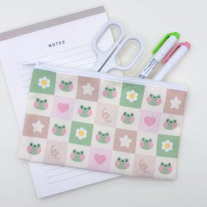 May include: A patterned pencil case featuring green frogs, flowers, stars, and hearts. White scissors and two highlighters are visible inside. A notepad with the word "NOTES" is also present. The case has a white zip.