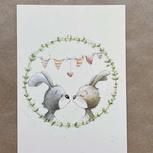 Könnte beinhalten: Eine Aquarell-Illustration von zwei Kaninchen, die sich in einem grünen Kranz küssen, über dem eine Girlande mit Flaggen hängt.