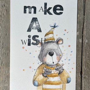 Könnte beinhalten: Eine Aquarell-Illustration eines grauen Bären, der einen gestreiften Partyhut trägt und auf einem weißen Hintergrund mit goldenen und blauen Konfetti einen Wunsch ausbläst. Der Text "make a wish" ist in Schwarz geschrieben.