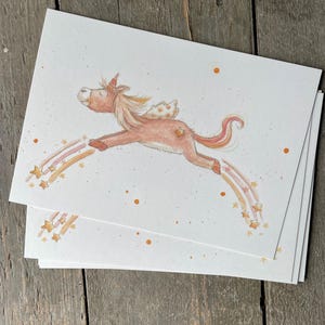 Könnte beinhalten: Eine Aquarell-Illustration eines braunen Einhorns mit goldenem Horn und Flügeln, das durch die Luft fliegt. Das Einhorn ist von rosa und orangefarbenen Regenbögen mit goldenen Sternen umgeben.