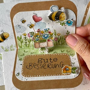 May include: A handmade get-well card with a watercolour illustration. The card shows a bouquet of flowers, bees, and the German phrase "Gute Besserung." The card is held by a person.