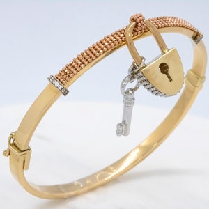 May include: A gold bangle bracelet with a lock and key charm. The bracelet is made of two-tone gold, with a rose gold band and a yellow gold lock and key charm. The lock and key charm is accented with small diamonds.