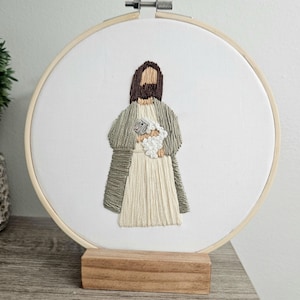 May include: Embroidered artwork in a wooden hoop depicts a figure holding a lamb. The figure is dressed in a cream-coloured robe and a green cloak. The lamb is white. The artwork is mounted on a wooden stand.
