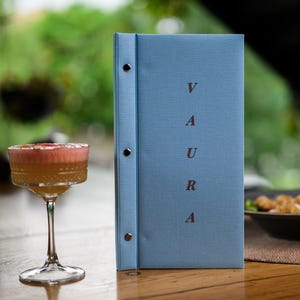 May include: A light blue vertical menu cover with the word VAURA printed in dark red letters, featuring a spine secured by three silver metal rivets. Beside it stands a stemmed glass filled with a golden liquid topped with pink foam.