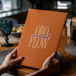 May include: A rectangular, burnt orange menu cover with a textured fabric finish. The front features the white text ORO and FLOW in a tall, elegant font, with the word cocktail written in a smaller, cursive, dark purple script in between.