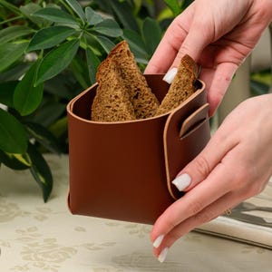 May include: A square, brown leather storage container with rounded corners and a folded, overlapping side panel. The container holds three slices of dark brown, textured bread. The material has a smooth finish with visible light-coloured edges.