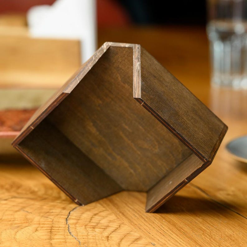 Side view of a dark walnut-stained plywood angled napkin holder with white paper napkins on a white clean surface. The open angled top, dark finish, and clean construction are shown from the side profile. Handcrafted custom engraved wooden napkin holder for pub table service, restaurant dining, bar counters, and hospitality interior styling with personalized laser engraving and plywood stain color options on Etsy marketplace.