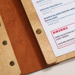 Natural plywood cover open on a table, interior pages with "Drinks" section visible. Leather spine on the right edge. This is a drinks menu specific shot — same plywood format as the food menu but showing the drinks section. Useful for buyers planning matching covers for food and drinks menus. Having the same plywood cover format across both menus creates a consistent table presentation — a detail that communicates care and professionalism in the dining room without being expensive to achieve.