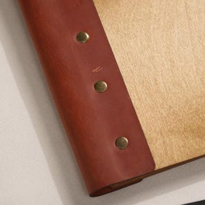 Close-up of the corner area where the plywood surface meets the leather binding strip. Silver rivet is visible. The join between the plywood and leather is clean — the leather sits flush against the wood surface with no gaps. For a product that combines plywood and leather, the quality of the join between them is a reliable indicator of the overall build standard. A clean, flat join suggests consistent manufacturing; a raised edge or gap would raise questions about long-term durability in use.