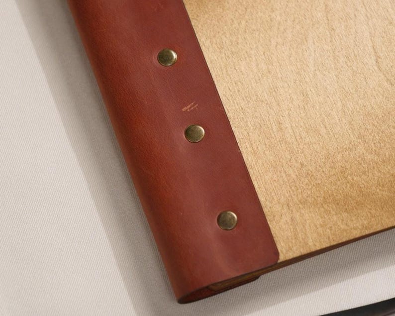 Close-up of the corner area where the plywood surface meets the leather binding strip. Silver rivet is visible. The join between the plywood and leather is clean — the leather sits flush against the wood surface with no gaps. For a product that combines plywood and leather, the quality of the join between them is a reliable indicator of the overall build standard. A clean, flat join suggests consistent manufacturing; a raised edge or gap would raise questions about long-term durability in use.