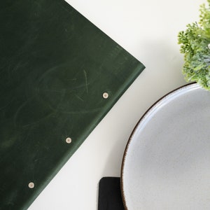 Dark forest green Crazy Horse leather menu cover with custom embossed logo standing upright on a modern white restaurant table next to gold cutlery and a white ceramic plate. Front view lifestyle product photo highlighting the genuine aged leather texture, personalized branding and snap button closure. Designed for cafes, bistros and upscale fine dining restaurants requiring elegant branded menu solutions.