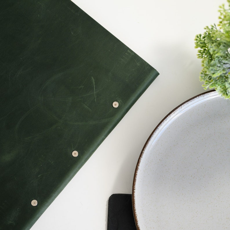 Dark forest green Crazy Horse leather menu cover with custom embossed logo standing upright on a modern white restaurant table next to gold cutlery and a white ceramic plate. Front view lifestyle product photo highlighting the genuine aged leather texture, personalized branding and snap button closure. Designed for cafes, bistros and upscale fine dining restaurants requiring elegant branded menu solutions.