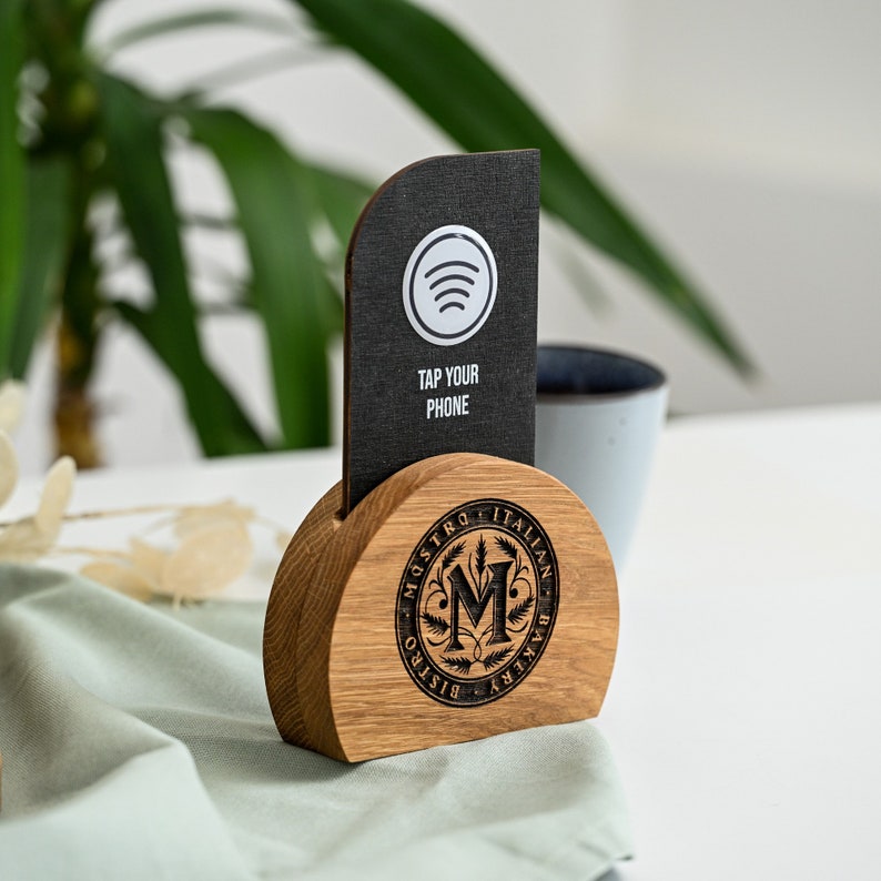 Side angle view of a custom NFC contactless menu stand showing the slim black display panel with Tap Your Phone text and the natural oak semicircle base with Maestro Italian restaurant logo engraved on the wood surface. The elegant combination of black panel and warm oak base creates a sophisticated table accessory for Italian restaurants, wine bars and modern dining venues offering NFC-based contactless digital menu access.