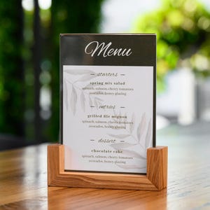 May include: A wooden menu stand with a clear acrylic sign that says "Menu" and lists the menu items: starters, spring mix salad, entrees, grilled file mignon, and dessert, chocolate cake. The menu items all include spinach, salmon, cherry tomatoes, avocados, and honey glazing.
