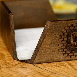 Wide lifestyle shot of a dark walnut-stained plywood napkin holder with engraved pub logo and white napkins on a wooden bar table with glassware and food dishes visible in the background in a real pub interior. The branded wooden napkin holder is shown in an authentic bar dining atmosphere. Custom engraved wooden napkin holder for pub table service, craft beer bar dining, restaurant interiors, and personalized hospitality woodcraft branding.