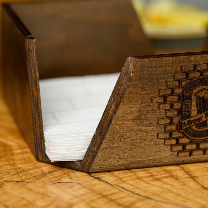 Wide lifestyle shot of a dark walnut-stained plywood napkin holder with engraved pub logo and white napkins on a wooden bar table with glassware and food dishes visible in the background in a real pub interior. The branded wooden napkin holder is shown in an authentic bar dining atmosphere. Custom engraved wooden napkin holder for pub table service, craft beer bar dining, restaurant interiors, and personalized hospitality woodcraft branding.