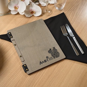 a square grey stone-textured menu cover with "Moi" in clean script burned into the front. Metal hinge hardware on the corners and spine gives it an industrial edge that contrasts well with the soft grey surface. Gold cutlery on the wooden table in the background. The overall look is calm and precise — Japanese minimalist aesthetics applied to a restaurant product. The stone texture is matte and cool, different from the warm wood and leather options elsewhere in the range.