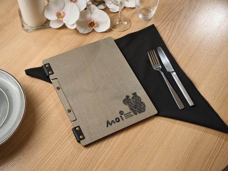 a square grey stone-textured menu cover with "Moi" in clean script burned into the front. Metal hinge hardware on the corners and spine gives it an industrial edge that contrasts well with the soft grey surface. Gold cutlery on the wooden table in the background. The overall look is calm and precise — Japanese minimalist aesthetics applied to a restaurant product. The stone texture is matte and cool, different from the warm wood and leather options elsewhere in the range.