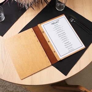 Natural plywood cover lying open on a table, interior pages showing printed menu content. Leather spine on the right edge, silver rivets along the outer edge. The table gives a different perspective from the shots in the listing. Interior pages are well-loaded and lying flat. This shot is more about atmosphere than technical detail — the circular table, warm light, and open plywood cover together create a welcoming, relaxed dining impression that communicates the product's intended environment.