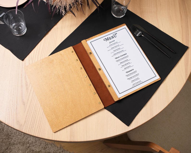 Natural plywood cover lying open on a table, interior pages showing printed menu content. Leather spine on the right edge, silver rivets along the outer edge. The table gives a different perspective from the shots in the listing. Interior pages are well-loaded and lying flat. This shot is more about atmosphere than technical detail — the circular table, warm light, and open plywood cover together create a welcoming, relaxed dining impression that communicates the product's intended environment.