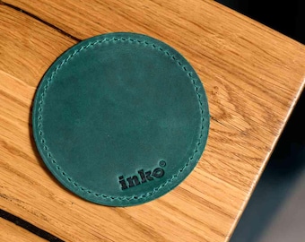 May include: A round, teal leather coaster with stitched edges. The coaster has the word "inko" printed in black near the bottom. The coaster is sitting on a wooden surface.