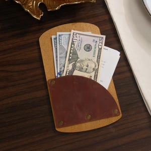 May include: A tan leather bill holder with a brown leather pocket, holding a stack of U.S. currency and a receipt. The bill holder is on a dark wooden surface. The pocket is secured with brass rivets.