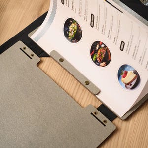 Overhead flat lay of the Mori cover open on a wooden table. Circular food photography on the interior pages. Metal hinge hardware. White ceramic plates and wooden-handled cutlery alongside. This overhead composition is the most editorial of all the product shots — it looks like something from a food magazine rather than a product listing. That's not a bad thing for a listing — it communicates that the cover belongs in an environment where design and presentation are taken seriously.