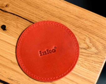 May include: A round, red leather coaster with stitched edges. The coaster has the embossed word "inko" in the center. The coaster is on a wooden surface.