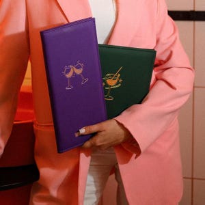 May include: A purple wine list holder with a gold illustration of two clinking wine glasses. A green wine list holder is behind the purple one. The word "WIN" is visible at the bottom of the purple holder.