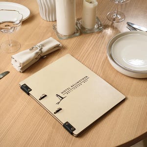 May include: A restaurant table setting featuring a wooden menu with the text "HOTEL BELLWETHER BELLINGHAM BAY", plates, silverware, a wine glass, and candles. The menu is light-colored with black accents and a black leather corner. The table is made of light wood.