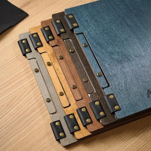Close-up of a dark denim blue square menu cover showing the metal hinge hardware on the corner and spine in detail. The blue textured surface is visible, the hinge mechanism clearly articulated. This is a quality assurance shot focused entirely on the hardware — showing it's well-made and properly aligned. The corner hinge is a distinctive design feature that sets these covers apart from the standard rivet or snap-button closure styles common across the rest of the product range.