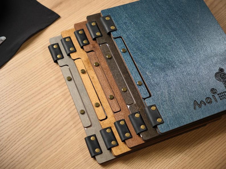 Close-up of a dark denim blue square menu cover showing the metal hinge hardware on the corner and spine in detail. The blue textured surface is visible, the hinge mechanism clearly articulated. This is a quality assurance shot focused entirely on the hardware — showing it's well-made and properly aligned. The corner hinge is a distinctive design feature that sets these covers apart from the standard rivet or snap-button closure styles common across the rest of the product range.