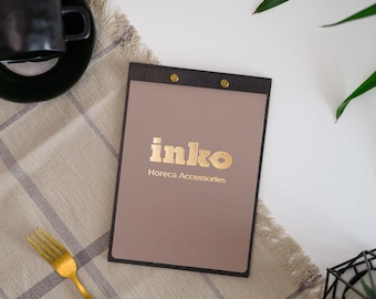 Puede incluir: Un portamenús con el texto dorado "inko Horeca Accessories" sobre un fondo topo. El soporte está enmarcado en negro y asegurado con remaches dorados. También se ven un tenedor dorado y una taza de café negra.