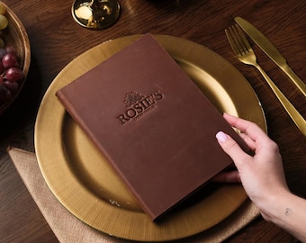 May include: A brown leather bound menu with the text "Rosie's,  Gourmet Gallery" embossed on the front. The menu is on a gold charger plate.