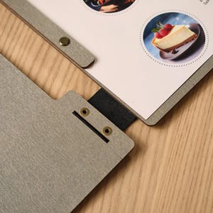 Grey wood-textured square Moi cover open on a wooden table, interior pages showing circular food photography images and minimal menu text. Metal hinge on the spine. White ceramic plate alongside. The circular food photos inside contrast interestingly with the square format of the cover — a deliberate design choice in the mock-up that gives the interior a gallery-like quality. Clean, considered, and very on-brand for a venue with a strong visual identity.