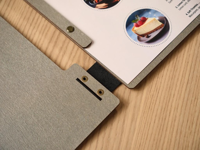 Grey wood-textured square Moi cover open on a wooden table, interior pages showing circular food photography images and minimal menu text. Metal hinge on the spine. White ceramic plate alongside. The circular food photos inside contrast interestingly with the square format of the cover — a deliberate design choice in the mock-up that gives the interior a gallery-like quality. Clean, considered, and very on-brand for a venue with a strong visual identity.