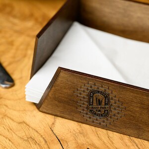 Close-up angled view of a dark walnut-stained plywood napkin holder with white paper napkins and laser-engraved pub roundel logo prominently displayed on the front panel. The precise engraving quality, dark surface contrast, and open-box construction are highlighted. Custom branding detail of a handcrafted wooden pub napkin holder for craft beer bars, restaurant table service, dining room interiors, and personalized hospitality woodcraft branding on Etsy.