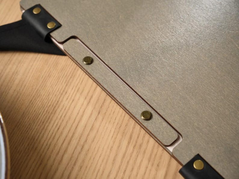 Grey wood-textured Mori cover alongside a white ceramic plate on a wooden table. Metal hinge hardware on the spine. Simple, clean composition. The white plate and grey cover create a very neutral, refined table setting. No cutlery, no props, just the cover and the plate. This minimalist styling choice suits the Mori brand aesthetic perfectly — Japanese design sensibility applied to the product photography as well as to the product itself.