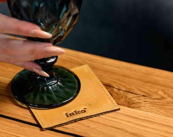 May include: A teal-colored, diamond-patterned glass with a matching base sits on a square, tan leather coaster. The coaster has the word "inko" embossed on it. The glass is held by a person with long nails. The coaster is on a wooden surface.