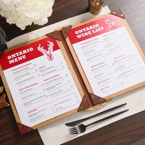 Peut inclure: Menus et cartes des vins ouverts avec du texte rouge et blanc sur un cadre en bois. Le menu indique "Ontario Menu" avec un logo de cerf. La carte des vins indique "Ontario Wine List" avec un logo de verre à vin. Une fourchette et un couteau sont sur la table.