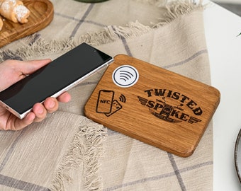 May include: A wooden serving board with the text "TWISTED SPOKE" and a motorcycle graphic. The board has a white sticker with a Wi-Fi symbol and an NFC symbol. A smartphone is held near the board.
