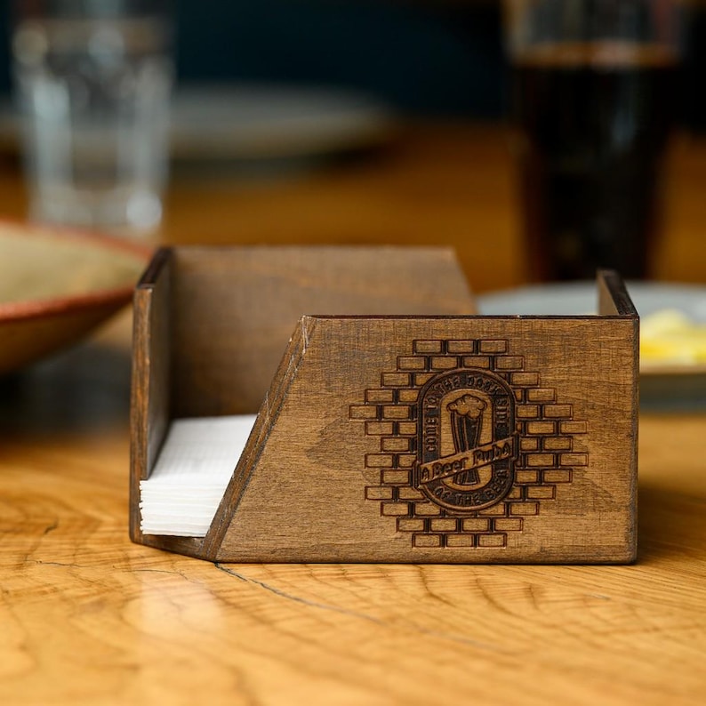 Handcrafted dark walnut-stained plywood open angled napkin holder with laser-engraved pub roundel logo on the front panel, holding white paper napkins on a real wooden bar table. The open-top angled construction and dark stained plywood finish are clearly visible in an authentic pub interior setting. Custom branded wooden napkin holder for pub table service, craft beer bars, restaurant dining, and hospitality interior branding with personalized laser engraved woodcraft.