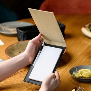 May include: A black leather server's notepad with a wooden cover, held open to reveal a blank white notepad and a silver pen. The notepad is on a wooden table, with plates and a basket in the background.