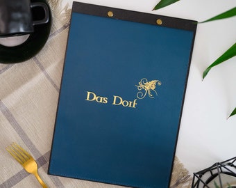 Puede incluir: Un portamenús rectangular de color azul oscuro con una banda superior negra sujeta por dos remaches dorados. La parte frontal presenta el texto Das Dorf en color dorado junto a un gráfico decorativo de espiral dorada.